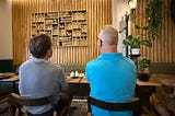 Two men are looking at the wooden art on the wall while seated by the dining table in the restaurant.