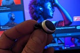 Foto eines modernen Hörgeräts vor einer Werbeanzeige die eine coole Person mit Hörgerät in einem musikalischen Event Setting zeigt