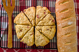 A wooden spatula sits on a metal rack next to freshly baked scones and a loaf of french bread. A red checked cloth is beneath them.