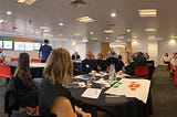Panoramic shot of a conference room with people sat in groups at round tables. There are flipchart sheets with post-its on along with other typical conference paraphenalia