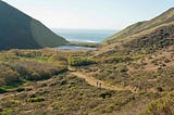 A sweeping landscape of the Marin Headlands, taken from the Tennessee Valley Trail, that includes hikers in the foreground and the Pacific Ocean in the distance.