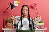 Relaxed Asian woman meditates at workplace, sits in zen pose against desktop, tries to relax after work, isolated over pink background