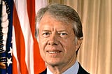 President Carter on an official portrait. Blue shirt, dark jacket, and regimental tie, the US flag on the background.