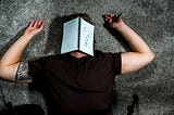 Man in black shirt and black tattoo on inner upper arm lieing on his back on a gray carpet, arms placed by his head with a notebook opened over his face and a big question mark drawn on one page.