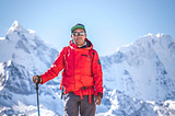 Kami Rita Sherpa stands in an orange jacket with a walking stick against the backdrop of the Himalayas.