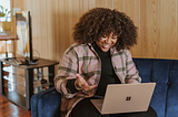 Photo by Surface on Unsplash — Woman with curly hair sitting on blue couch, smiling and gesturing to an open laptop computer on her lap