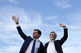 Secretary of Transportation Pete Buttigieg and his husband, Chasten, wave at the crowd framed against a blue sky.