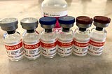 6 small, unopened peptide vials on a table with a bottle of bacteriostatic water behind them.