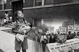 A black and white composite image showing a girl holding a baby and looking at the sky. Newspaper stating Dr. King Marches in Chicago with a rock on it. Protesters for fair housing.