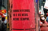 Lambe vermelho com a frase “sobrevivemos, eu e os meus. Desde sempre.” da Conceição Evaristo, colada num poste. Ao redor as pessoas erguem os punhos em meio a um protesto.