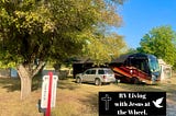 Big Red, our black and red Entegra Anthem motorhome, parked beneath shady trees at our RVICS Village home base in Smithville, Texas, with our site marker and RV Living with Jesus at the Wheel logo in the foreground on a peaceful sunny afternoon.