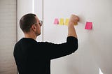 A man standing in front of a white wall with posted notes as he examines them.