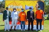 Group ohoto of two women and three men standing outside, wearing protective masks, and standing in front of a big and yellow photobooth where you can read “#Speak It Loud” with a woman holding a microphone