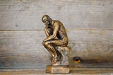 A brass man sculpture sitting with their hand on their chin slightly folded over, signifying a thinking pose.