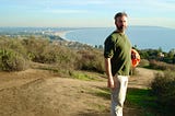 A young bearded man stands on a bluff overlooking a coastline