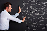 A photo of a man by a blackboard covered with many instances of the word “problems.”