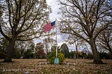A flagpole with an American flag, flanked by trees, in a cemetery on an overcast autumn day.