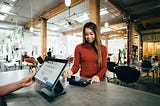 A woman with long hair swipes her credit card at a hair salon while someone behind the register operates a tablet device.