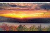 Photograph of the sun setting over a ridge on the far side of a body of water, with autumn-colors among the trees in the foreground. Photographer: David Dayton.