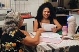 No canto esquerdo da imagem vemos a avó de Maysa sentada à mesa com sua neta, que sorri enquanto lê um dos textos escritos à mão pela avó. Elas estão sentadas na mesa da cozinha da avó de Maysa, em Coruripe.