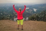 The author, a white woman with a long blonde braid, stands atop Mt. Lee in Los Angeles on an overcast day. Her back is to the camera and she faces the downtown skyline with her arms raised triumphantly above her head. In the foreground, the top edges of the Hollywood sign peek in.