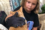 Young blonde girl using large glove to pet orange tabby cat