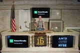 A white woman in a grey dress standing with a gavel, prentending to ring the closing bell at the New York Stock Exchange (NYSE). Monitors feature the TD Ameritrade Institutional logo.