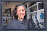 Portrait of author sitting at her desk with microphone in front of her, smiling at a camera, with her bookcase and paintings in the background