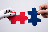 On the left is a robotic hand holding a red puzzle piece. On the right, a human hand holds a blue puzzle piece. The two hands are attempting to fit the puzzle pieces together.