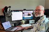 White man with a beard and red glasses smiles as he sits in front of a desk holding a laptop and two monitors.