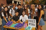 Smiling college students standing and sitting at a table covered with dental floss and condoms and a sign “FREE PFAS.”