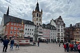 Picture of buildings in the city center of Trier, Germany. The buildings are constructed in traditional German architecture. There is a fountain on the left hand side of the square and there are people gathered around.