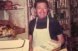Man wearing white apron at his grocery store, standing near scale
