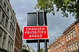 A red road sign with white text reads “CHANGED PRIORITIES AHEAD.” It is mounted on two black poles and stands in an urban street lined with historic brick and stone buildings. A tree with green leaves partially covers the top right corner of the image, while the sky is cloudy. The street appears quiet, with a few pedestrians and a car in the background.