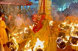 A performer surrounded by flaming torches during a nighttime ritual at a temple, with devotees and attendants watching him in awe.