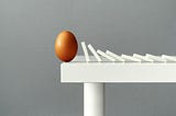 Dominos falling toward an egg that is upright at the edge of a table.