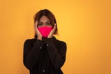 A woman wearing a long sleeved black button down shirt and a red face mask holds her hands to either side of her face and looks directly into the camera. She is posed against a dark yellow background.