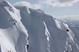 Picture of big mountains covered with untouched snow and mutliple cornices at the top. Some clouds in the sky, from far away. In the distance a skier is visible, the size of a tiny spec, coming down creating a seemingly impossible line.