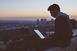 A man on his laptop, sitting on a wall atop a tall building. The backdrop is a city against an orange, darkening sky as the sun sets.