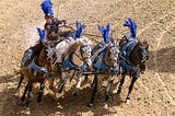 Photo of a young man in an Ancient Roman costume riding a chariot pulled by four horses. Both the horses and the driver are wearing blue feathers.