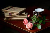 Poetry books set on top of one another next to a tea cup and two pink roses on a wooden table.