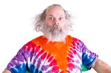 Senior man with grey hair and beard so surprised he’s wide-eyed, he is wearing a tie dye colorful t-shirt