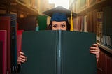 Woman in library wearing cap and gown looking out from a book she’s holding over the bottom half of her face