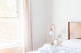 A light-filled bedroom with a bed with white sheets and a side table with a copper accent light and a plant.