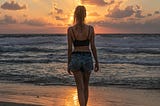 A woman in a black crop top and jean shorts watches the Sun set over the ocean on a beach.