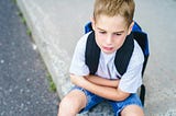 A little boy wearing a backpack sits on the sidewalk looking sad. His arms are loosely crossed in his lap.