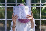 A woman in a pink hijab readinga book. She is holding the book in front of her face.