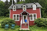 A 70 year old woman on the steps of her farmhouse-style red cottage near the woods.