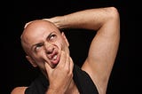 A bald man holds his head in disgust while he peers up. His other hand squeezes his mouth together causing pursed lips.