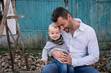 A male and a child boy sitting on a bench outside in front of a blue door in an autumn set up — Overprotective daddy making it through a sick child’s day